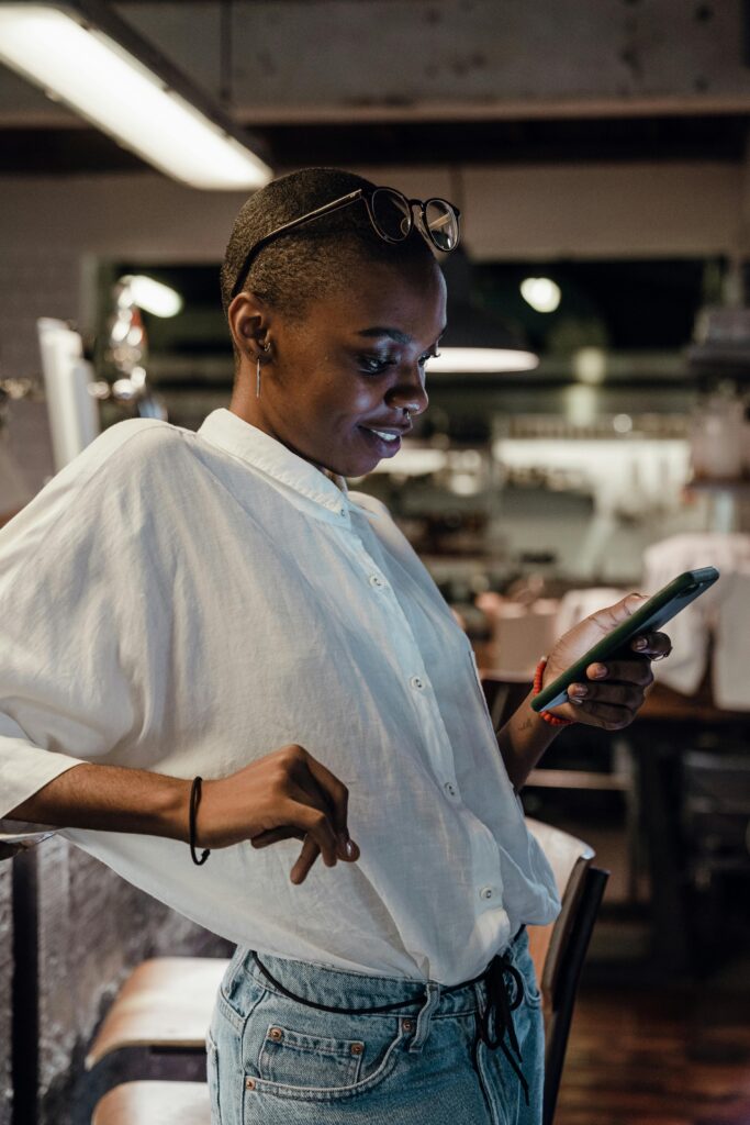 Young confident African American lady in trendy casual outfit with short hairstyle messaging on mobile phone and smiling lightly while leaning on bar counter