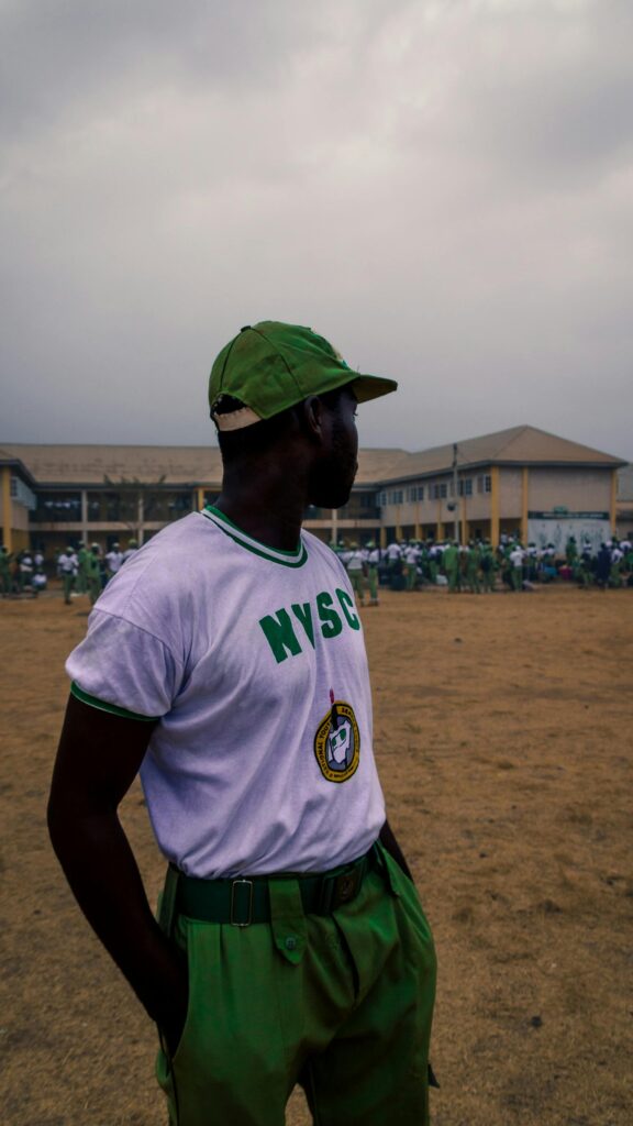 A person in NYSC uniform standing outside a building on a cloudy day.
